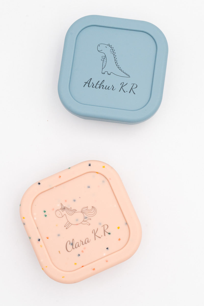 Deux boîtes moyennes, bleue et rose mouchetée, avec les prénoms Arthur K.R et Clara K.R et un dinosaure et une licorne gravés