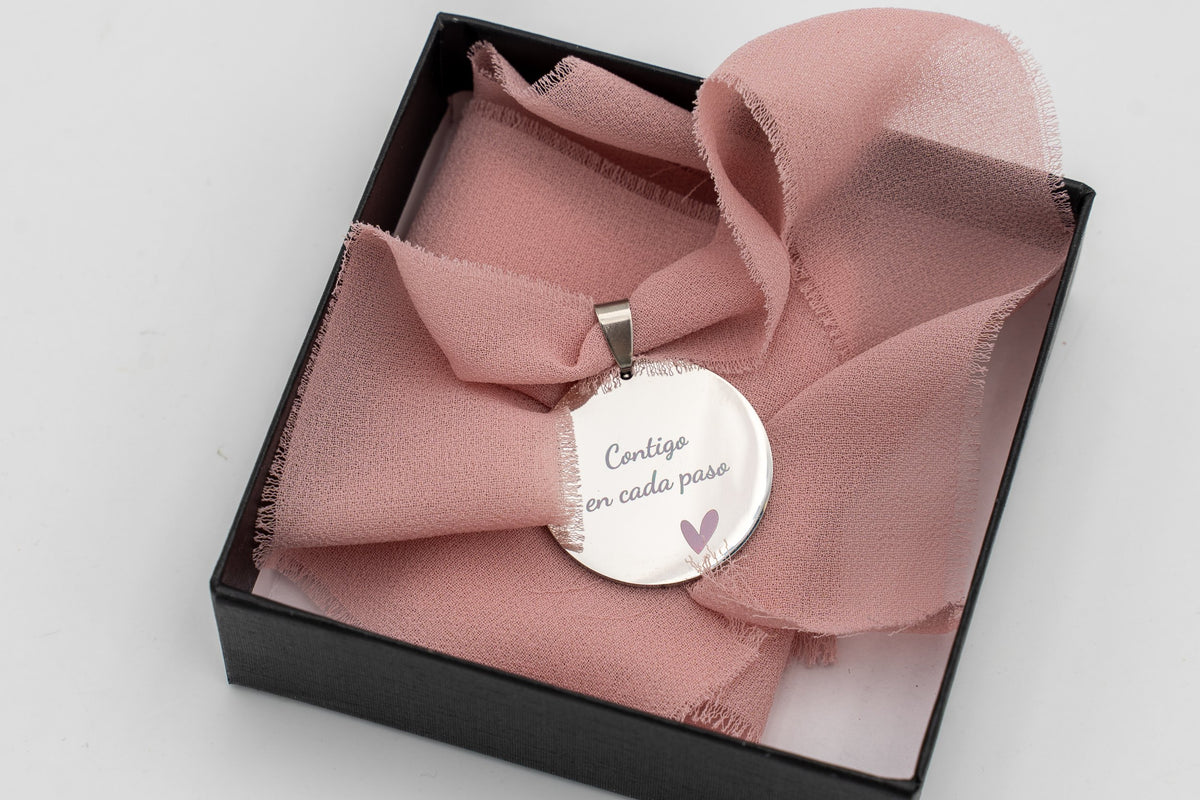 Médaille argentée avec la phrase « Avec toi à chaque pas » sur un ruban en mousseline rose, dans une boîte noire