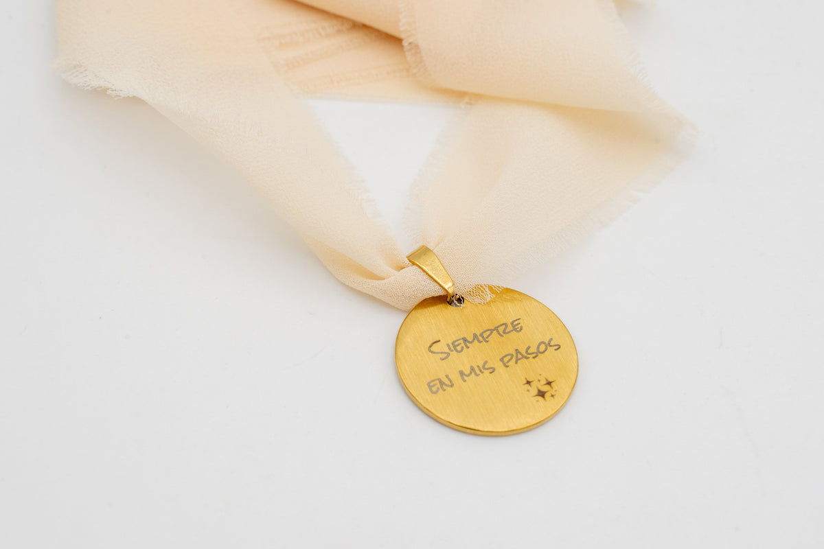 Médaille dorée avec la phrase « Toujours dans mes pas » nouée à un ruban en mousseline pêche