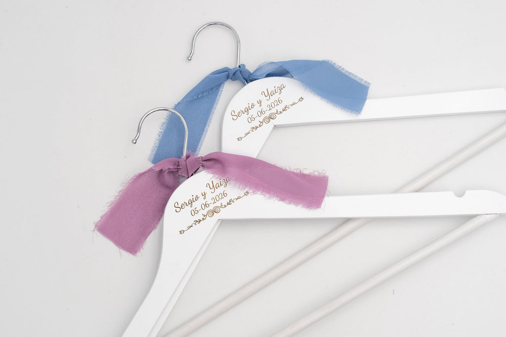 Deux cintres de mariage blancs avec les prénoms des mariés gravés et des nœuds en mousseline bleus et lilas