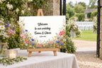 Panneau de bienvenue de mariage A3 en couleur, modèle Pradera, avec texte en anglais, sur un chevalet de table à l’entrée du jardin.